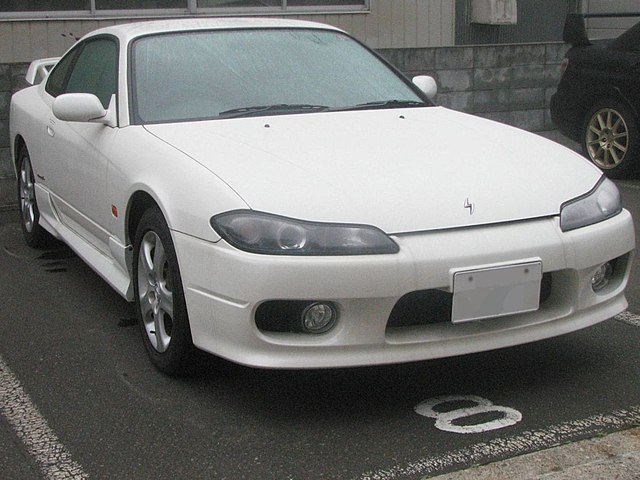 A 1999 Nissan Silvia S15 with a sleek, modern design, aggressive front bumper, and sporty rear wing, driving on a winding road with a sunset in the background.