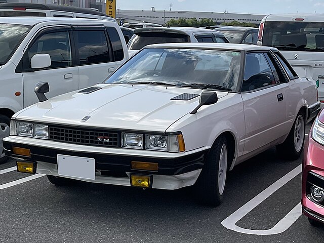 A 1980 Nissan Silvia S110 coupe with sharp-edged styling, pop-up headlights, and classic 80s design, parked in an urban setting.