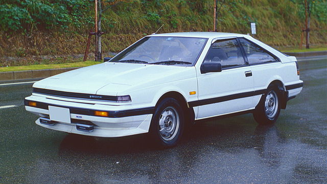  A 1984 Nissan Silvia S12 coupe with pop-up headlights, a sleek wedge-shaped design, and sporty alloy wheels, parked on a scenic mountain road.