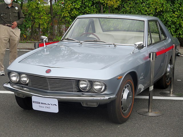 A classic 1965 Nissan Silvia CSP311 in pristine condition, featuring a sleek coupe design with chrome accents and round headlights, parked on a scenic road.