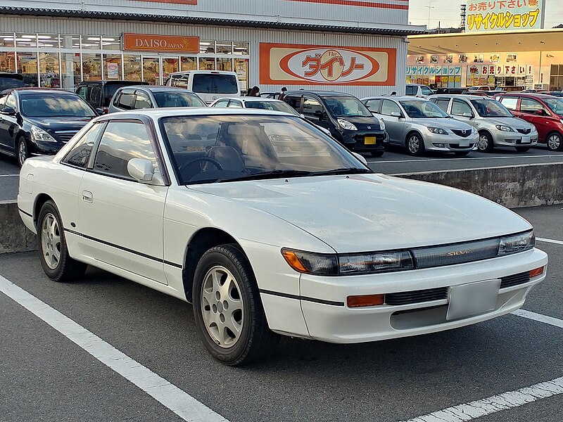 A 1989 Nissan Silvia S13 with a low-slung, aerodynamic design, pop-up headlights, and aftermarket wheels, drifting around a corner on a winding road.